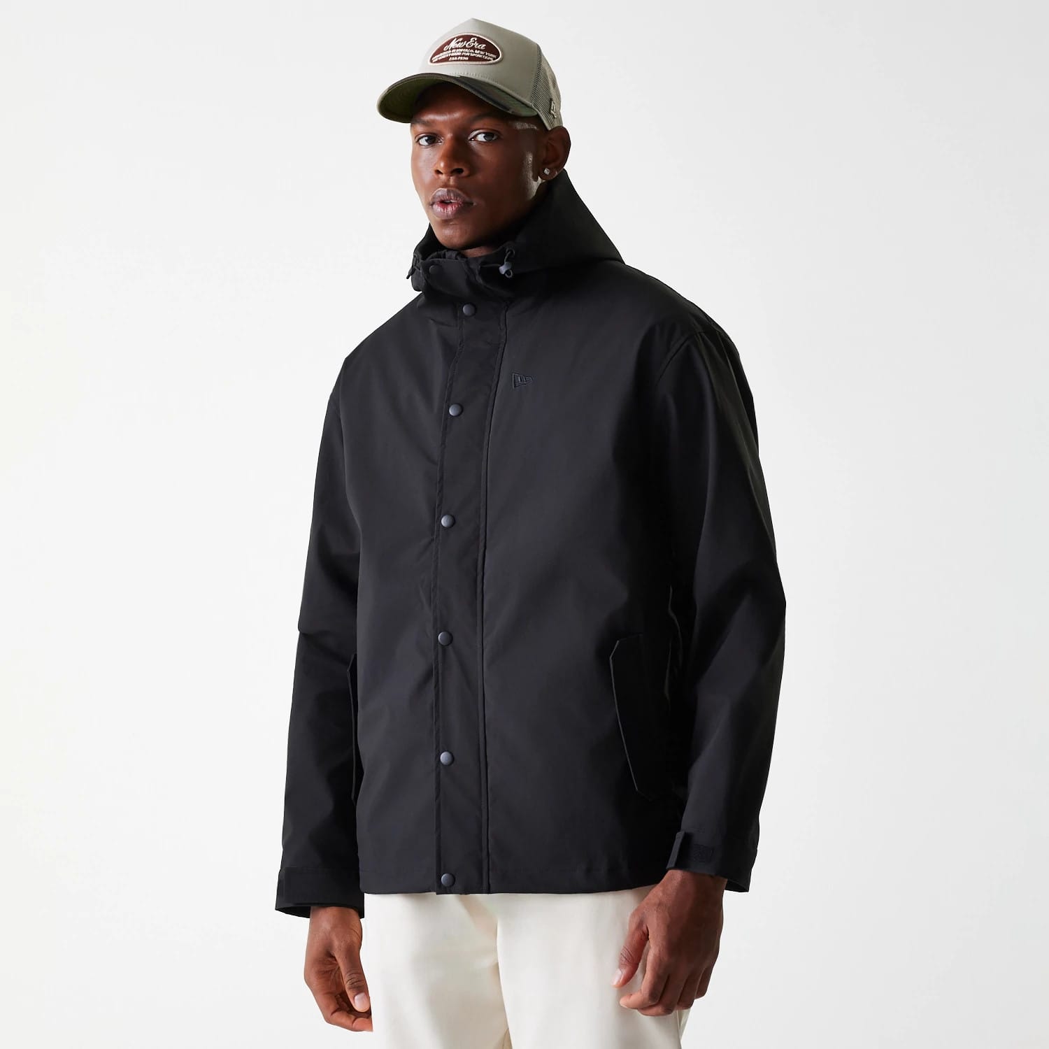 New Era Black Coach Jacket, on-model view showing a black button-up jacket with a raised New Era logo on the chest, a high collar and long sleeves, paired with a light grey cap displaying a contrasting patch on the front.