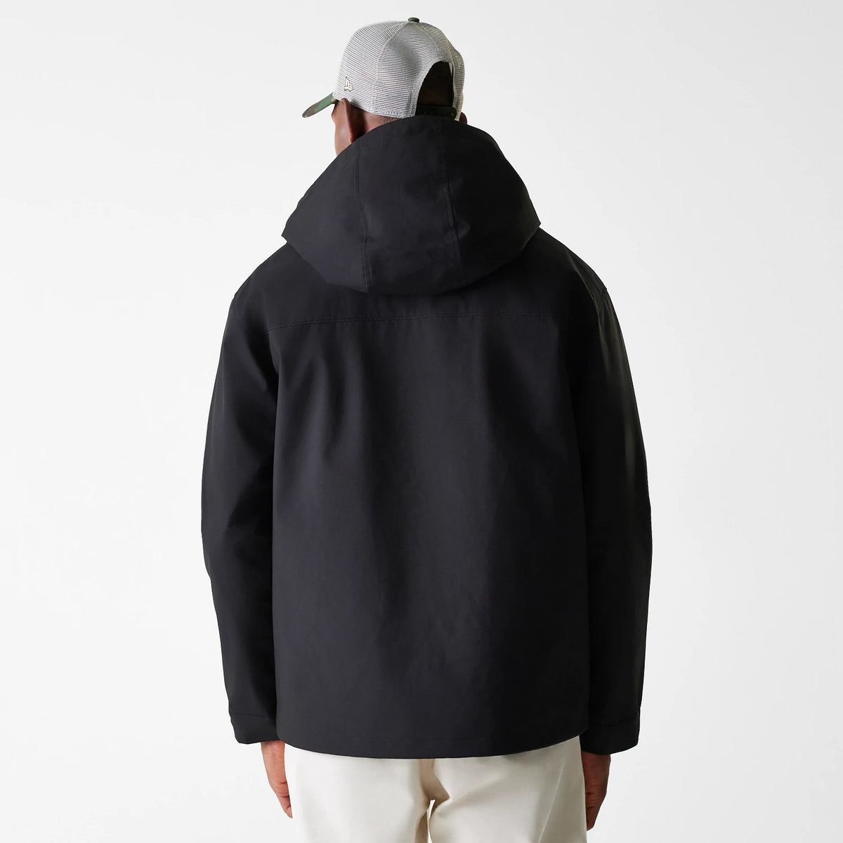 New Era Black Coach Jacket, rear view showing the plain black back with a large hood, the wearer dressed in light trousers and a grey mesh cap with a small embossed New Era logo on the side.