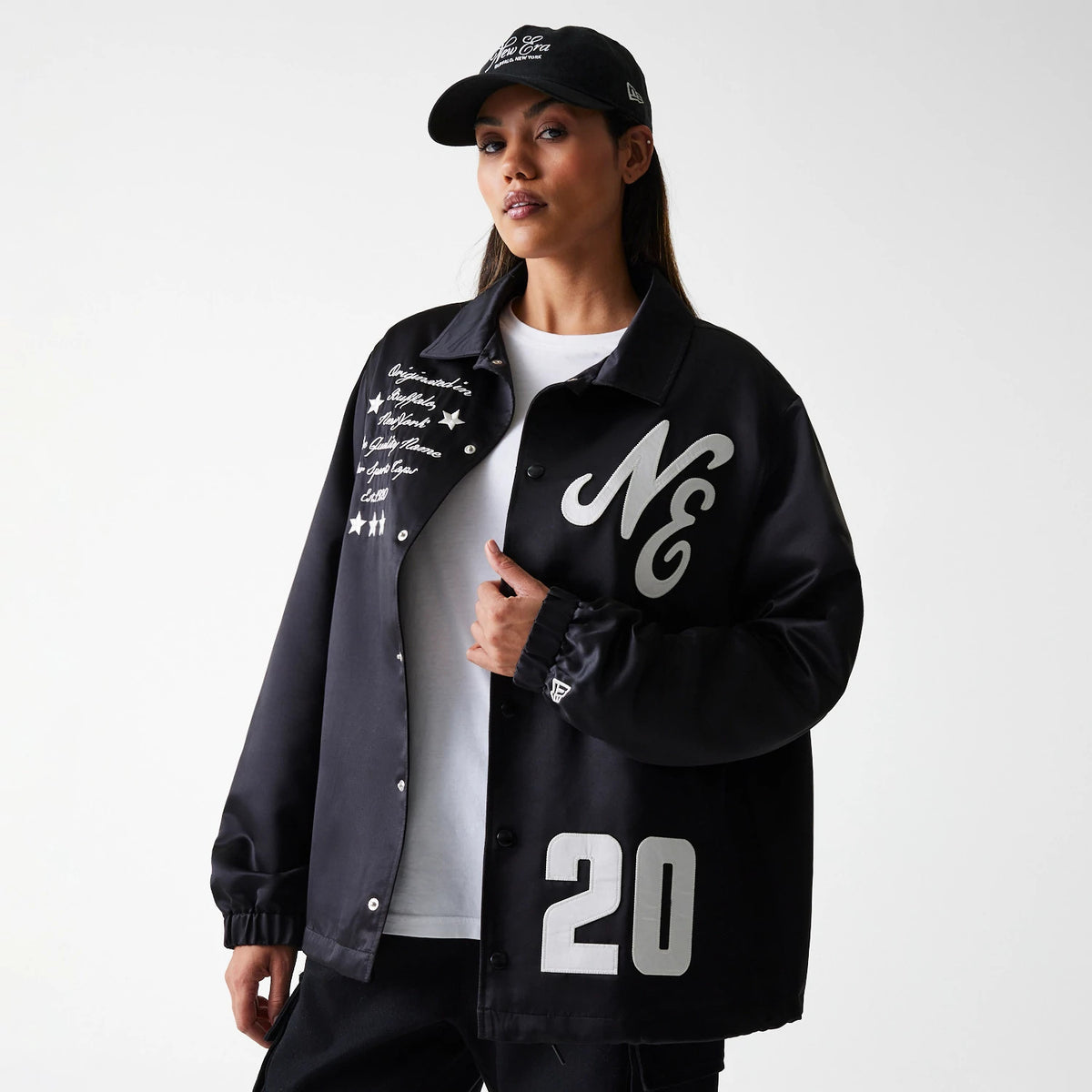 New Era Black Coaches Jacket, on-model view showing chest with a large 'NE' logo on the left side, embroidered script and stars on the right, and the number '20' at the hem, paired with a black cap.