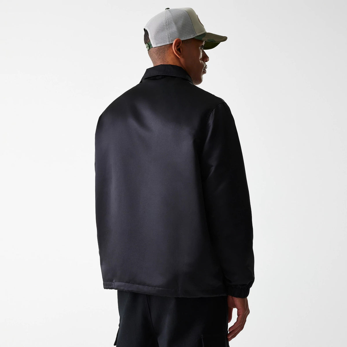 New Era Black Coaches Jacket, rear view showing the sleek black back panel extending from the collar down to the hem, paired with elasticated cuffs, worn by a person in a grey and camouflage cap.