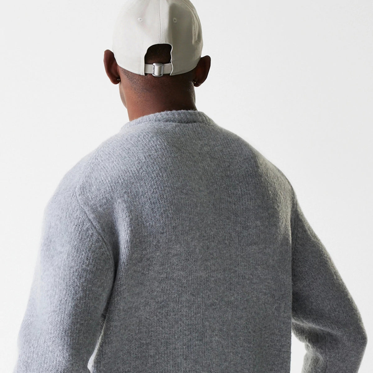 New Era Knit Grey Oversized Knit Sweater, back view on a model wearing a light-coloured cap, showing the textured knit pattern and relaxed fit of the sweater across the shoulders and upper arms.