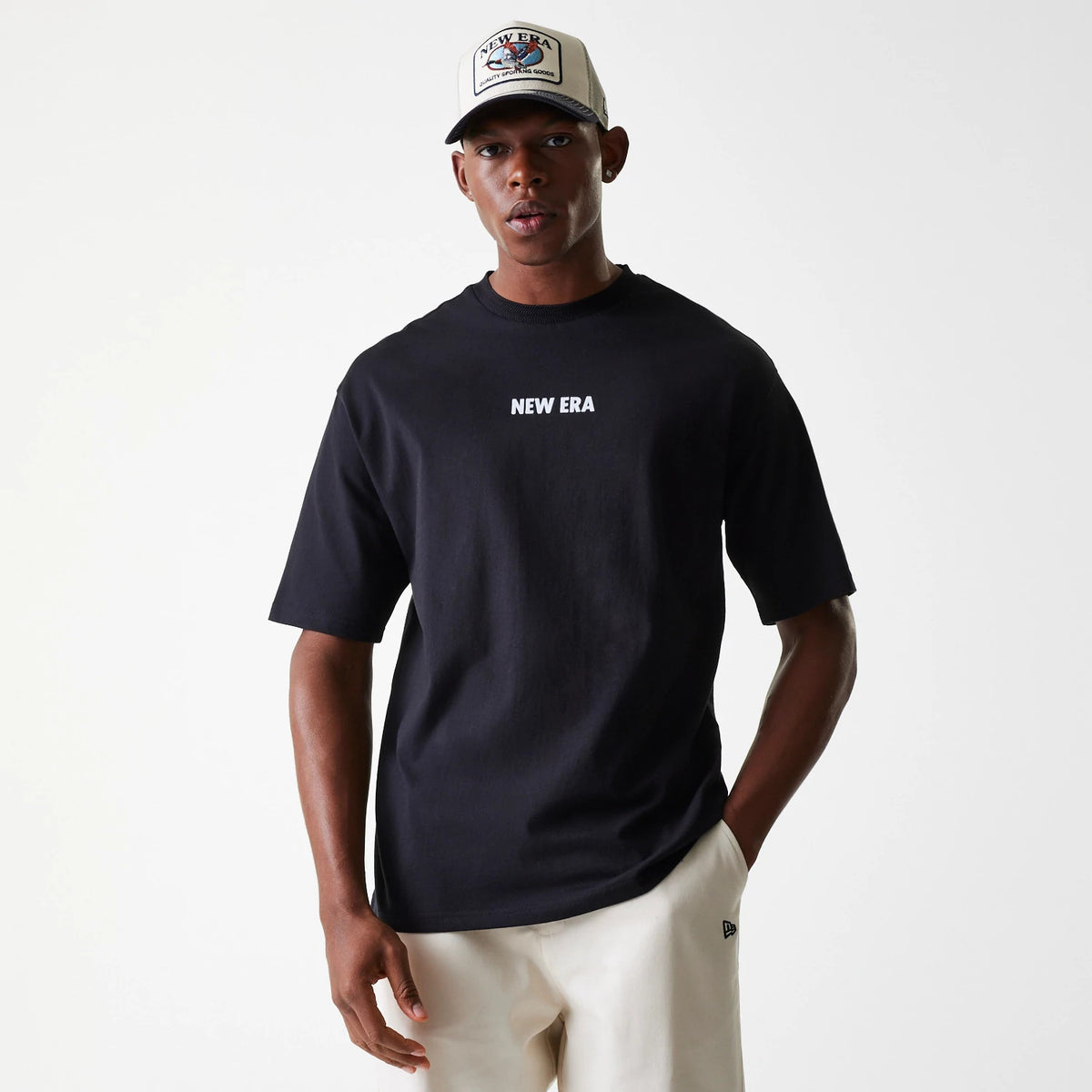 New Era Graphic Black Oversized T-Shirt, on-model view showing the black oversized fit with a white printed New Era logo centred on the chest and short sleeves, paired with a beige and black cap featuring a colourful patch on the front.