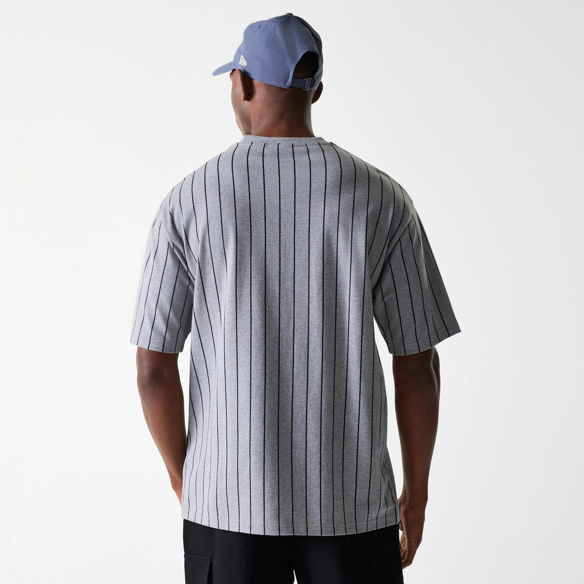 New Era Pinstripe Pastel Grey Oversized T-Shirt, on-model view showing the back with vertical black pinstripes on grey fabric, short sleeves and a crew neckline, paired with a man wearing a purple cap featuring a raised New Era logo on the side.