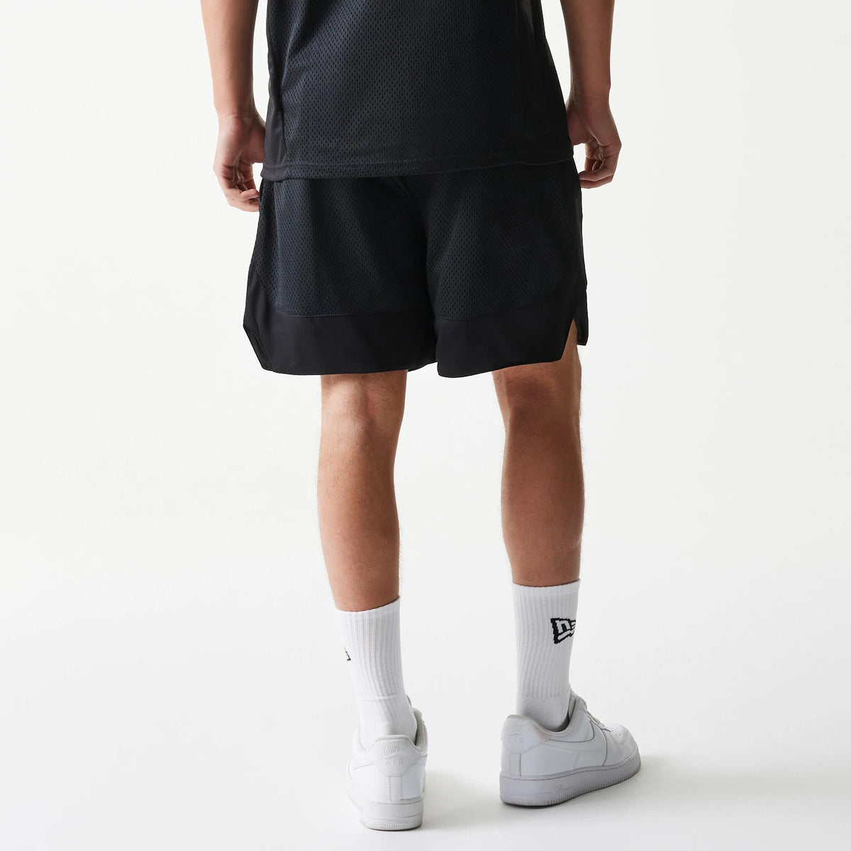 New Era Mesh Black Shorts, rear view showing the relaxed fit black shorts with a mesh texture, a plain waistband, and a black hem, worn on-model with white socks featuring a debossed New Era logo and white trainers.