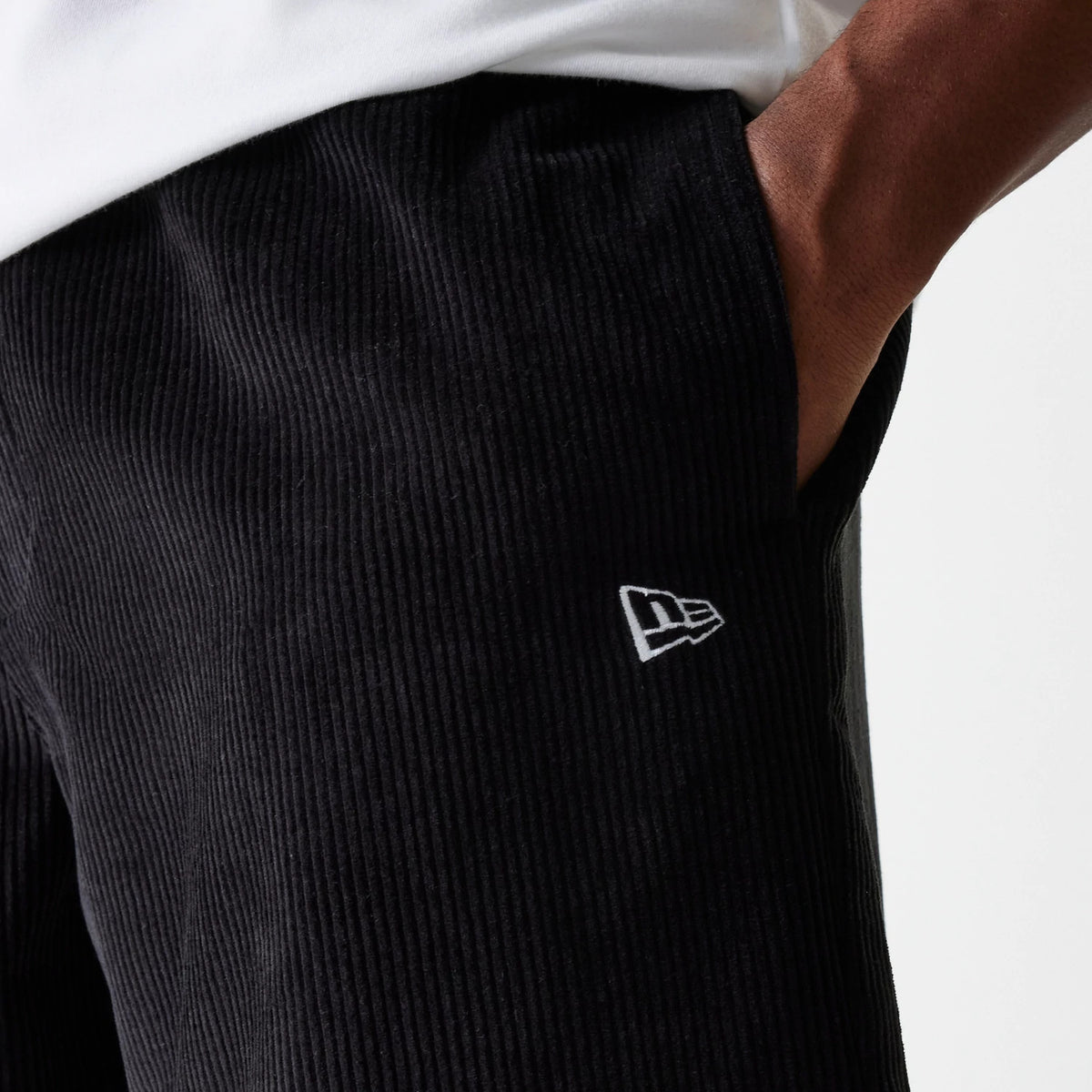 New Era Cord Black Shorts, on-model view showing the textured black surface with a small white embroidered New Era logo near the hem, positioned on the side of the shorts next to the pocket.