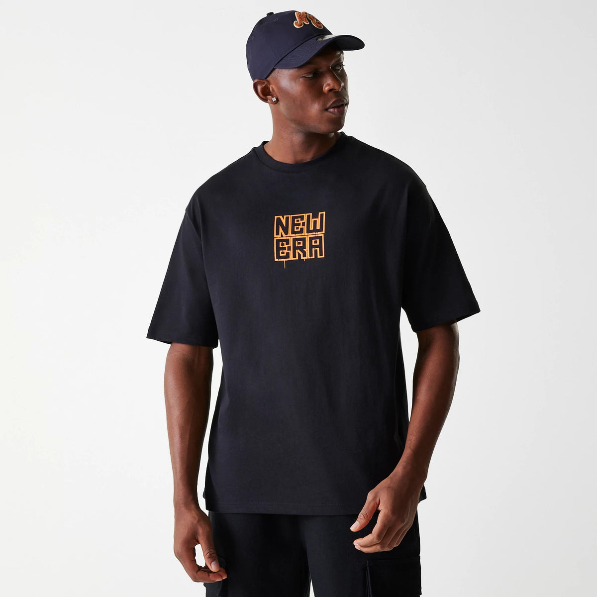 New Era Graffiti Graphic Black Oversized T-Shirt, on-model view showing the black t-shirt with an orange graffiti-style New Era logo centred on the chest, paired with a navy cap featuring an embossed logo.