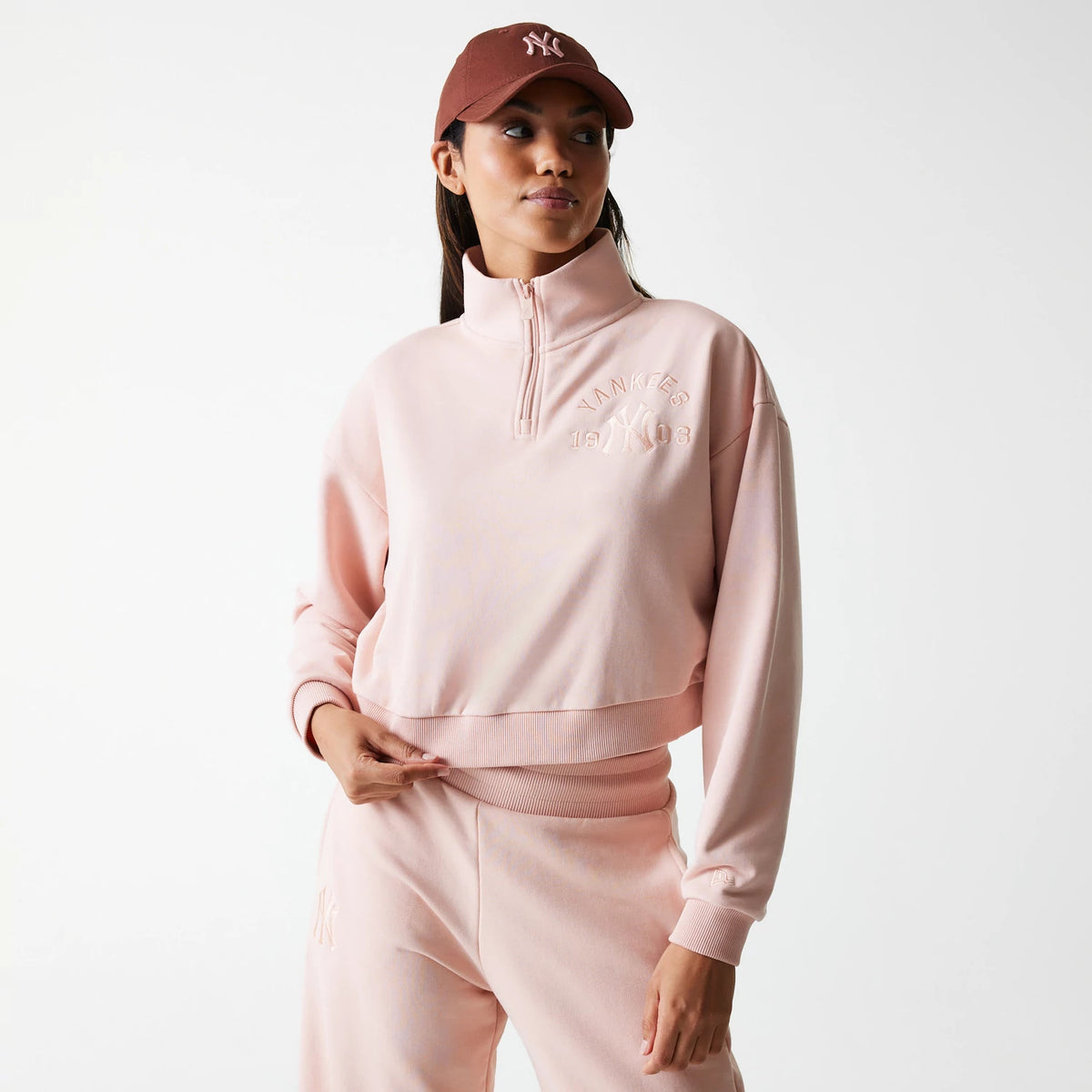 New Era New York Yankees Womens MLB Tonal Pastel Pink Cropped 1/4 Zip Sweater, on-model front view with raised embroidered Yankees logo across the chest, combined with a matching pastel pink tracksuit and a brown Yankees cap.