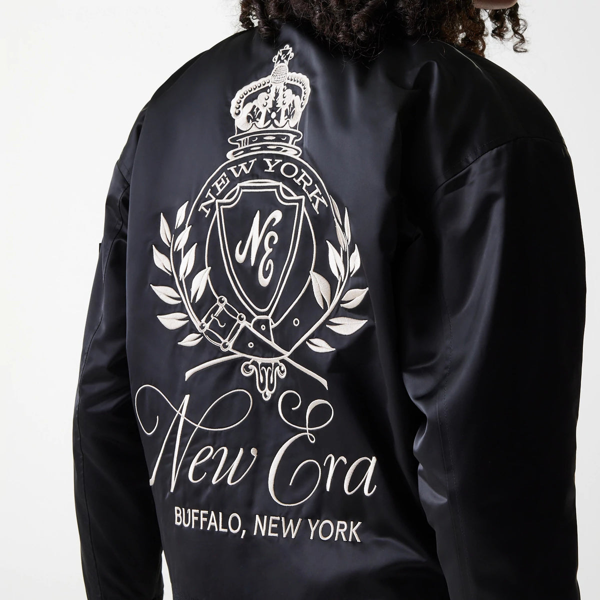 Womens New Era Black Satin Bomber Jacket, back view showing a large white embroidered crest design with New York and New Era text, along with flourishes and Buffalo, New York at the bottom of the black satin fabric.