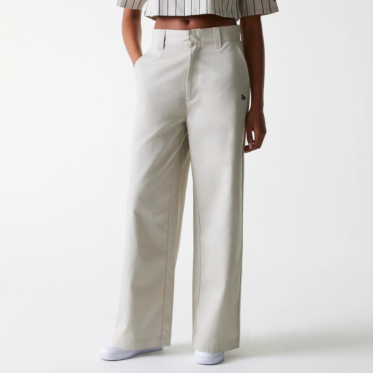 Womens New Era Light Beige Wide Leg Chino Trousers, front view on a model wearing a striped crop top, featuring side pockets, belt loops, and the New Era flag logo embroidered in black on the left thigh.