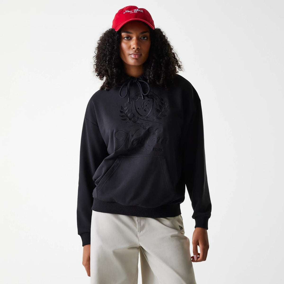 Womens New Era Crest Black Pullover Hoodie, front view showing the black hoodie with a large embossed crest and New Era logo on the chest, drawstrings hanging from the hood and a kangaroo pocket at the front, worn by a model with a red cap.