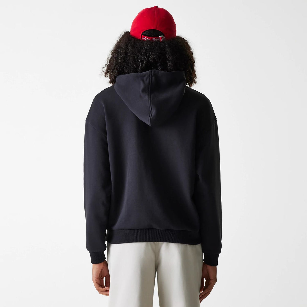 Womens New Era Crest Black Pullover Hoodie, rear view showing the relaxed black hoodie with a generous hood, the hem and cuffs ribbed for fit, worn by a model with a red cap, showcasing a casual look.