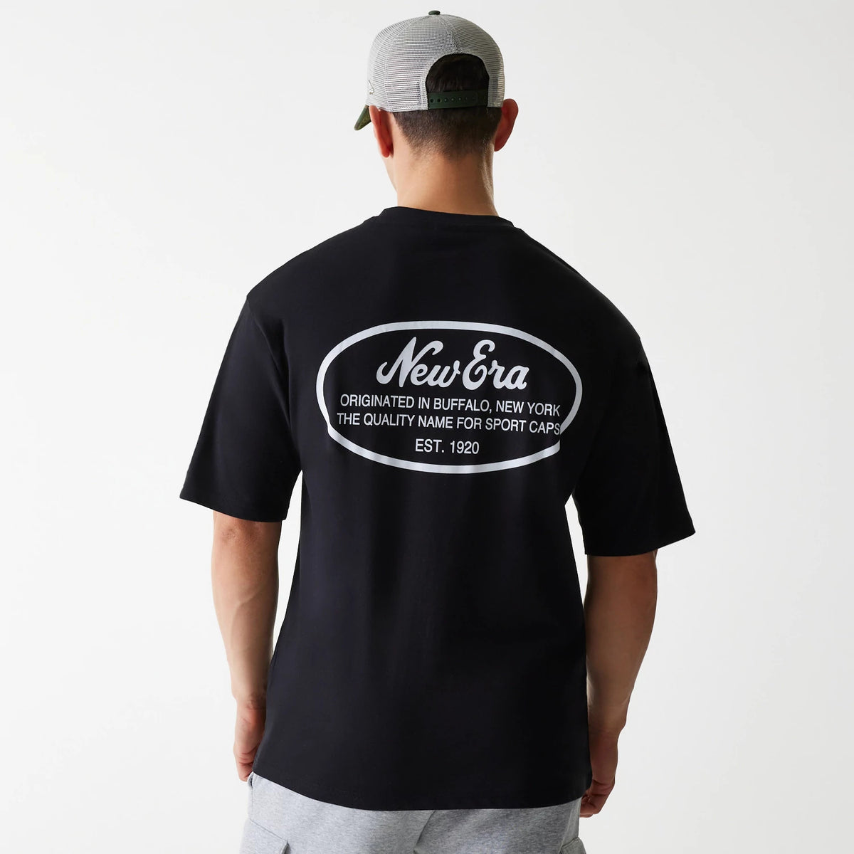 New Era Trucker Patch Black Oversized T-Shirt, rear view showing a large oval graphic with debossed New Era branding and text centred on the upper back, worn by a model in light grey shorts and a mesh-back baseball cap.