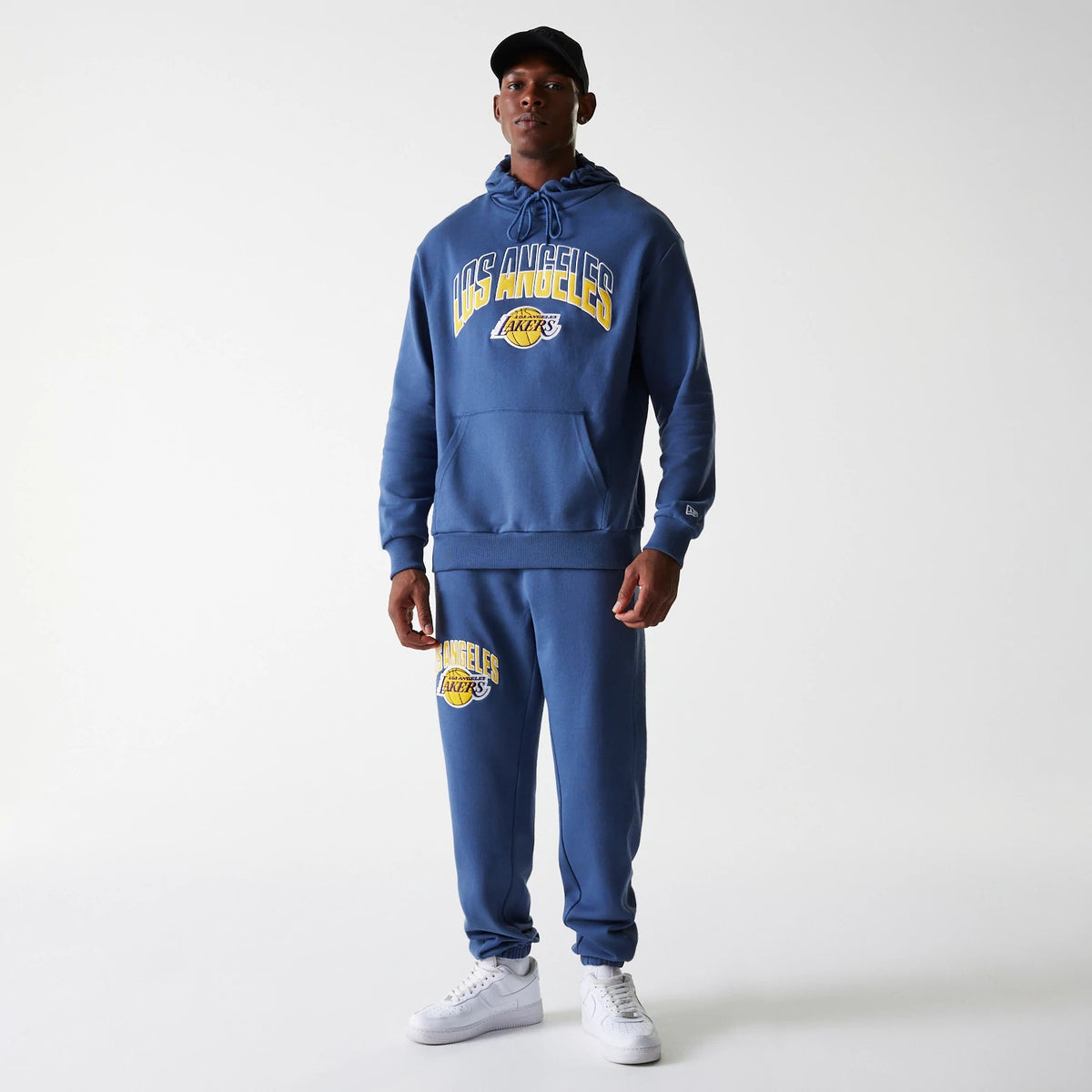 New Era LA Lakers NBA Arch Graphic Dark Blue Joggers, on a model with a matching hoodie, showing the LA Lakers logo embroidered in yellow and white on the left thigh, paired with white trainers and a black cap.