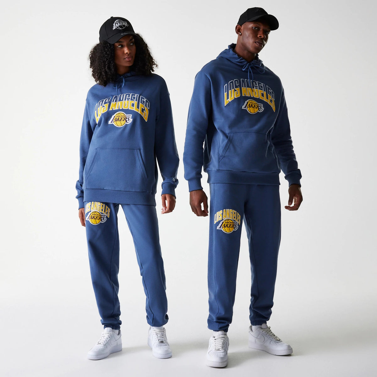 New Era LA Lakers NBA Arch Graphic Dark Blue Joggers, on a model paired with matching hoodie and cap, featuring the LA Lakers logo and lettering in yellow and white across the front of the left leg.