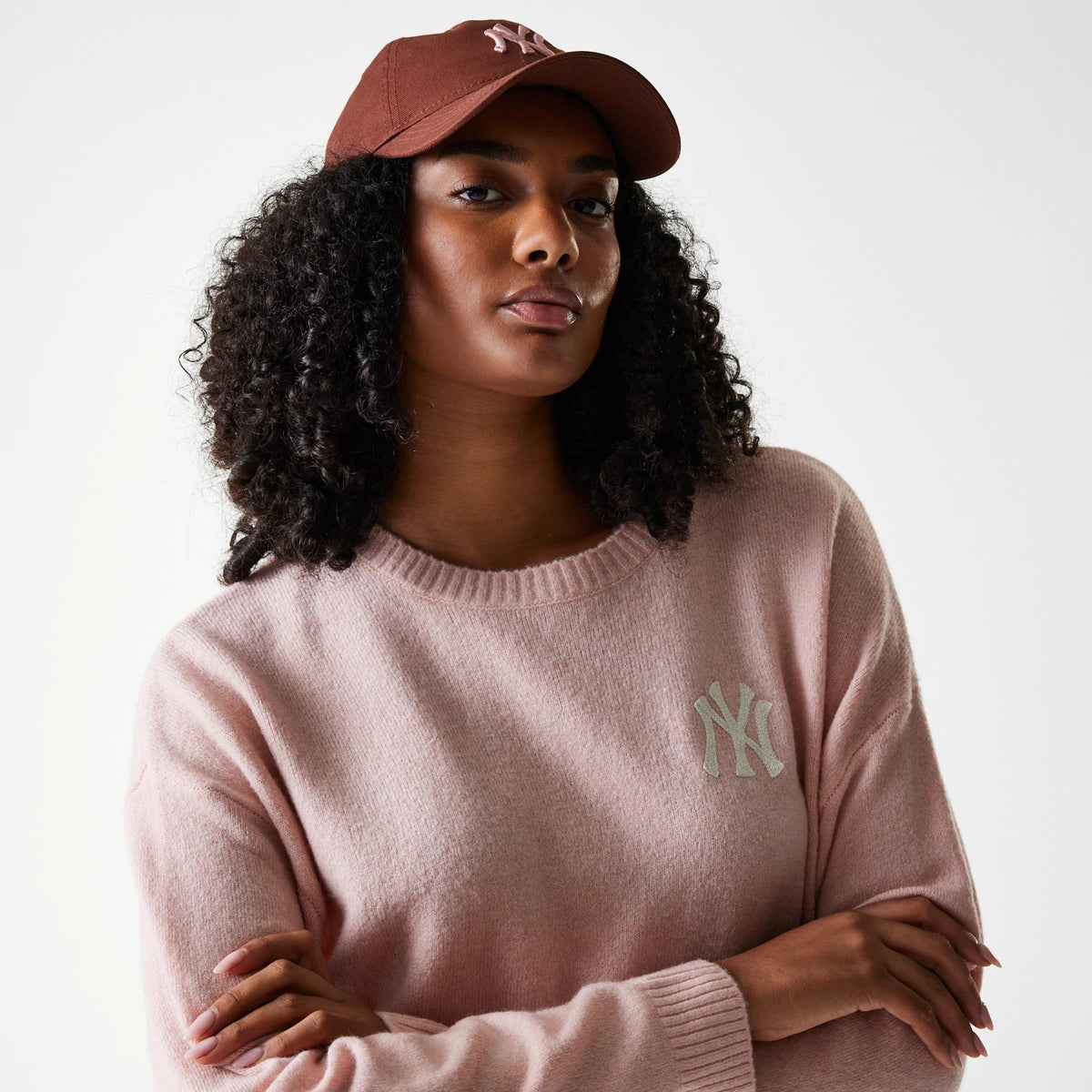 New Era New York Yankees Womens MLB Knit Pastel Pink Crew Neck Sweater, worn by a model with a brown cap, featuring the grey NY logo on the left chest and a ribbed neckline for a relaxed fit.