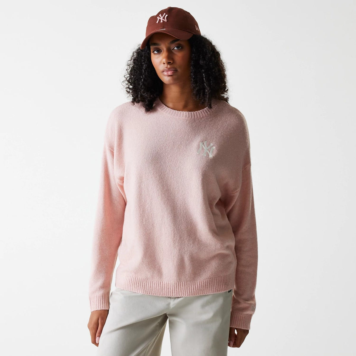 New Era New York Yankees Womens MLB Knit Pastel Pink Crew Neck Sweater, on-model view styled with a brown cap, showing the white embroidered NY logo on the left chest of the soft pastel pink sweater.