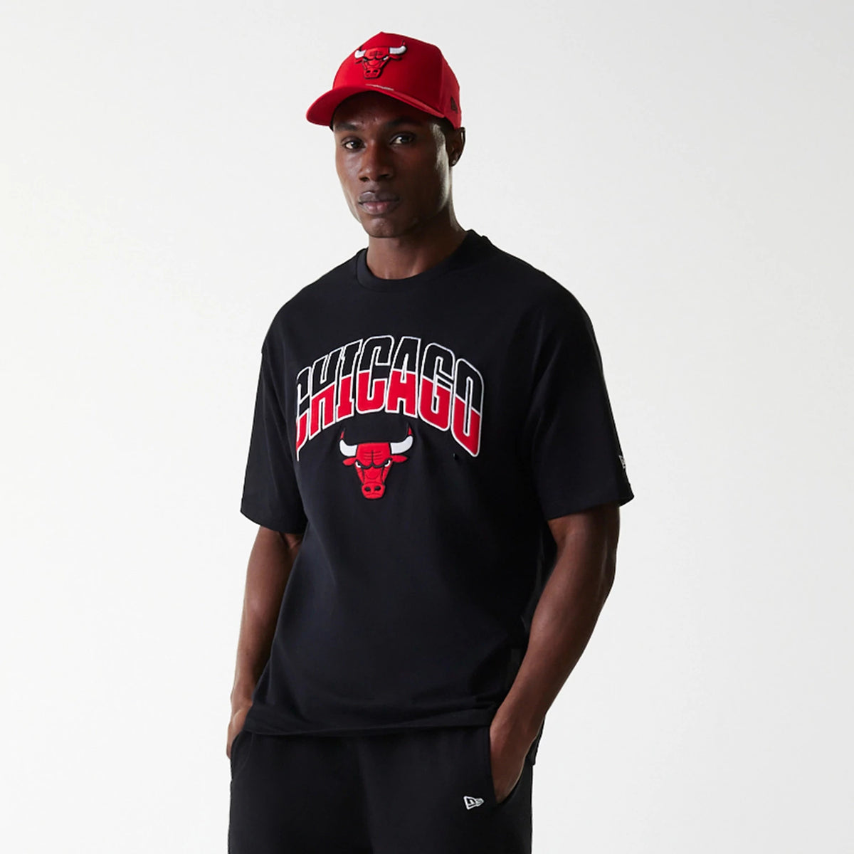 New Era Chicago Bulls NBA Arch Graphic Black Oversized T-Shirt, on-model view showing the large red and white CHICAGO text with a Bulls logo below across the chest, paired with a red cap featuring an embroidered Bulls logo on the front.