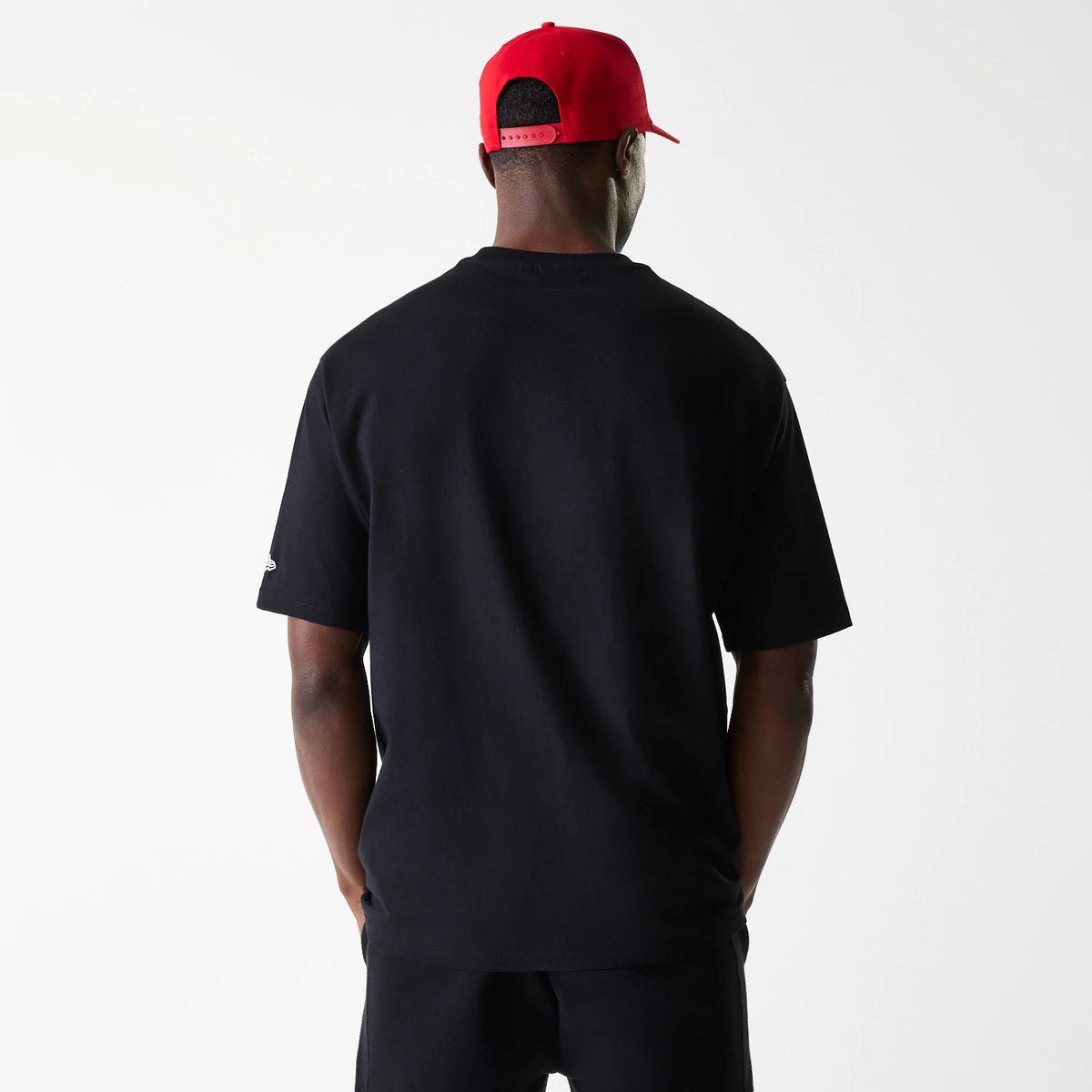 New Era Chicago Bulls NBA Arch Graphic Black Oversized T-Shirt, rear view showing the plain oversized black back, short sleeves, and a debossed New Era logo on the left sleeve, worn by a person in a red cap with a row of eyelets.