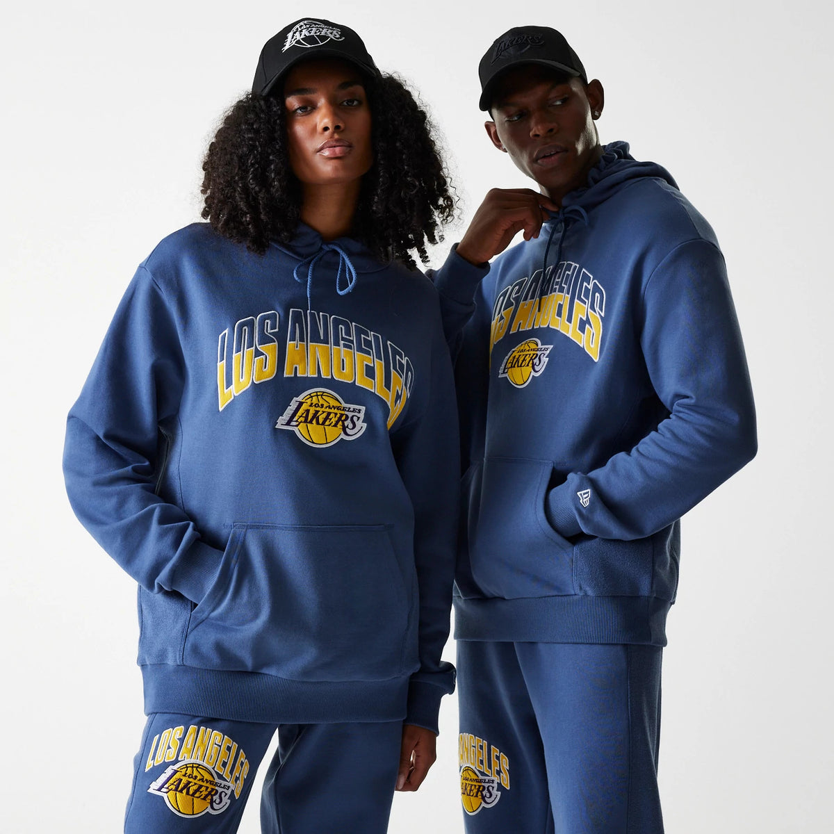 New Era LA Lakers NBA Arch Graphic Dark Blue Oversized Pullover Hoodie, on-model view showing two hoodies with Los Angeles Lakers embroidered on the chest, the models wearing black caps with a raised Lakers logo.