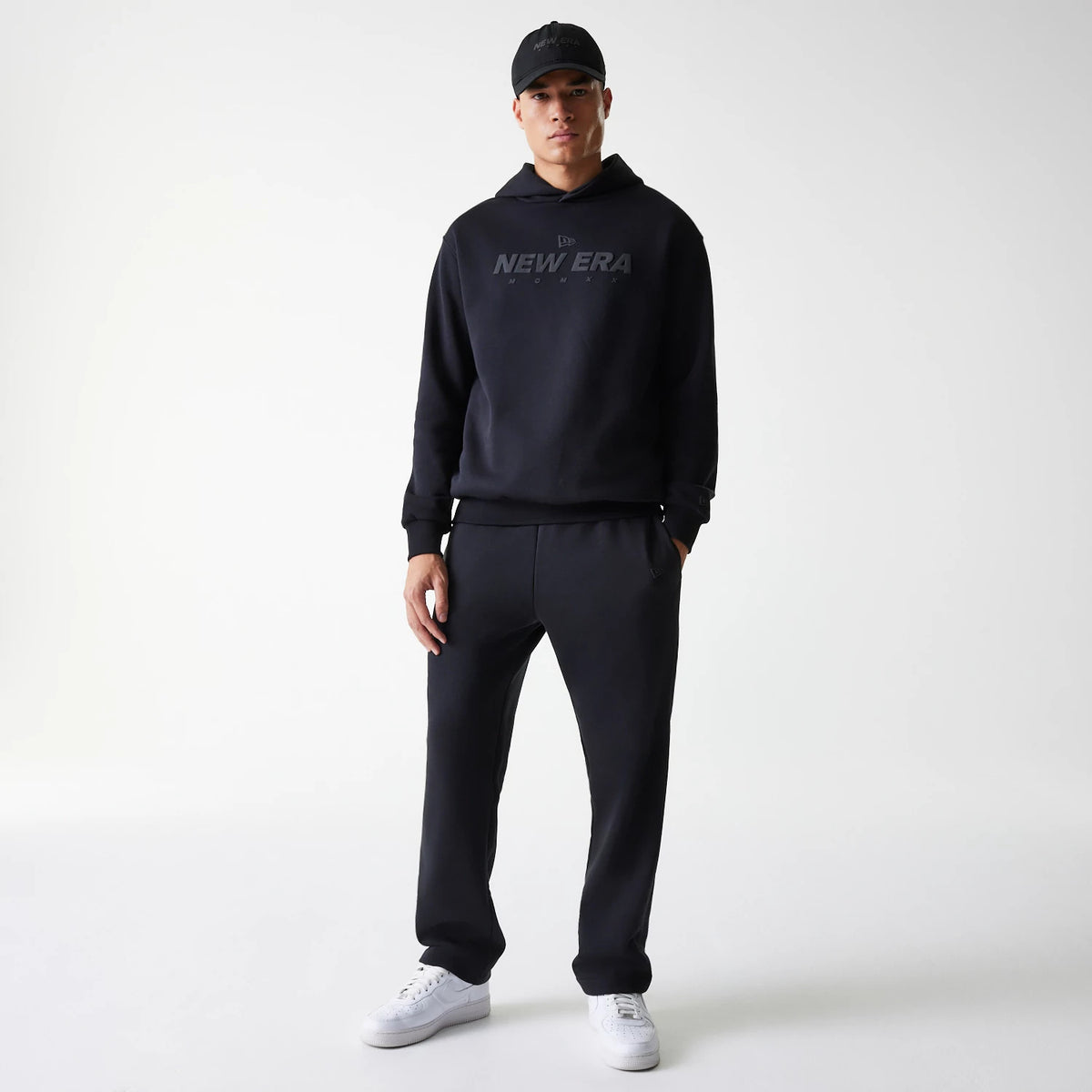 New Era Tech Black on Black Oversized Pullover Hoodie, on-model view showing the oversized black hoodie with a raised New Era logo on the chest, paired with black trousers and a black cap featuring the same raised logo.