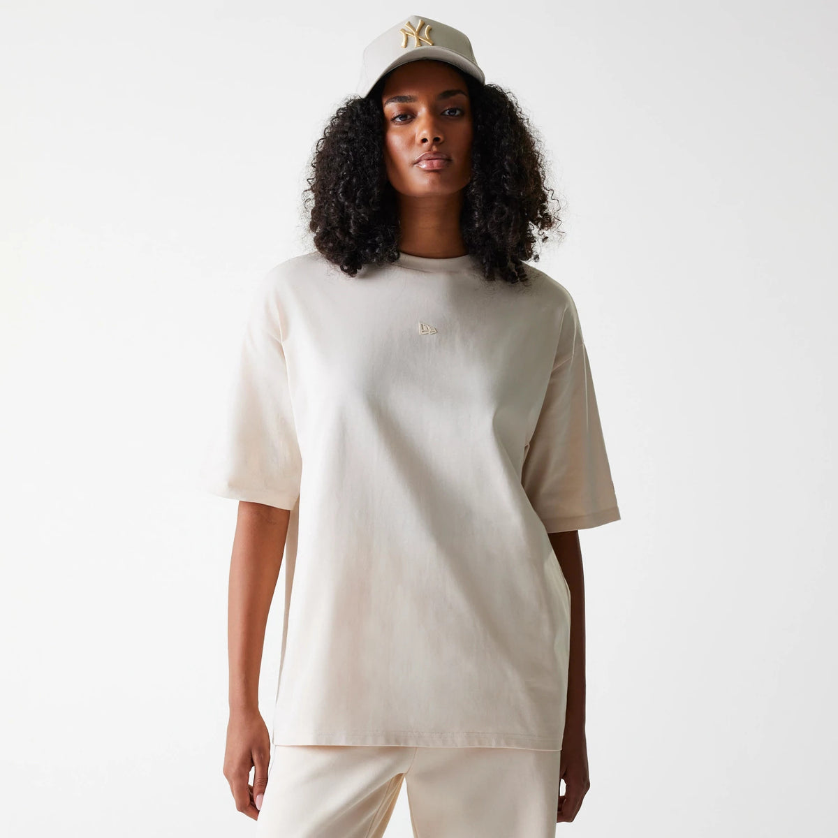 New Era Tech Light Beige Oversized T-Shirt, on-model view showing the light beige oversized fit with short sleeves, a raised New Era logo on the upper chest, worn with matching light beige trousers and a cap featuring a raised logo.