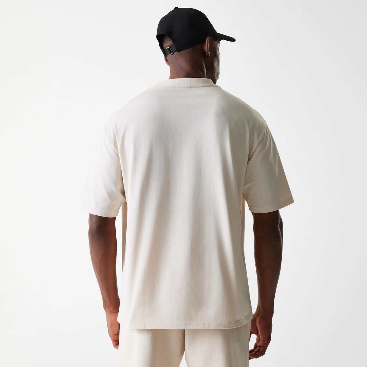 New Era Tech Light Beige Oversized T-Shirt, back view showing the plain light beige fabric with a relaxed fit on the shoulders and sleeves, worn by a person in matching shorts, with a black cap featuring an adjustable strap.