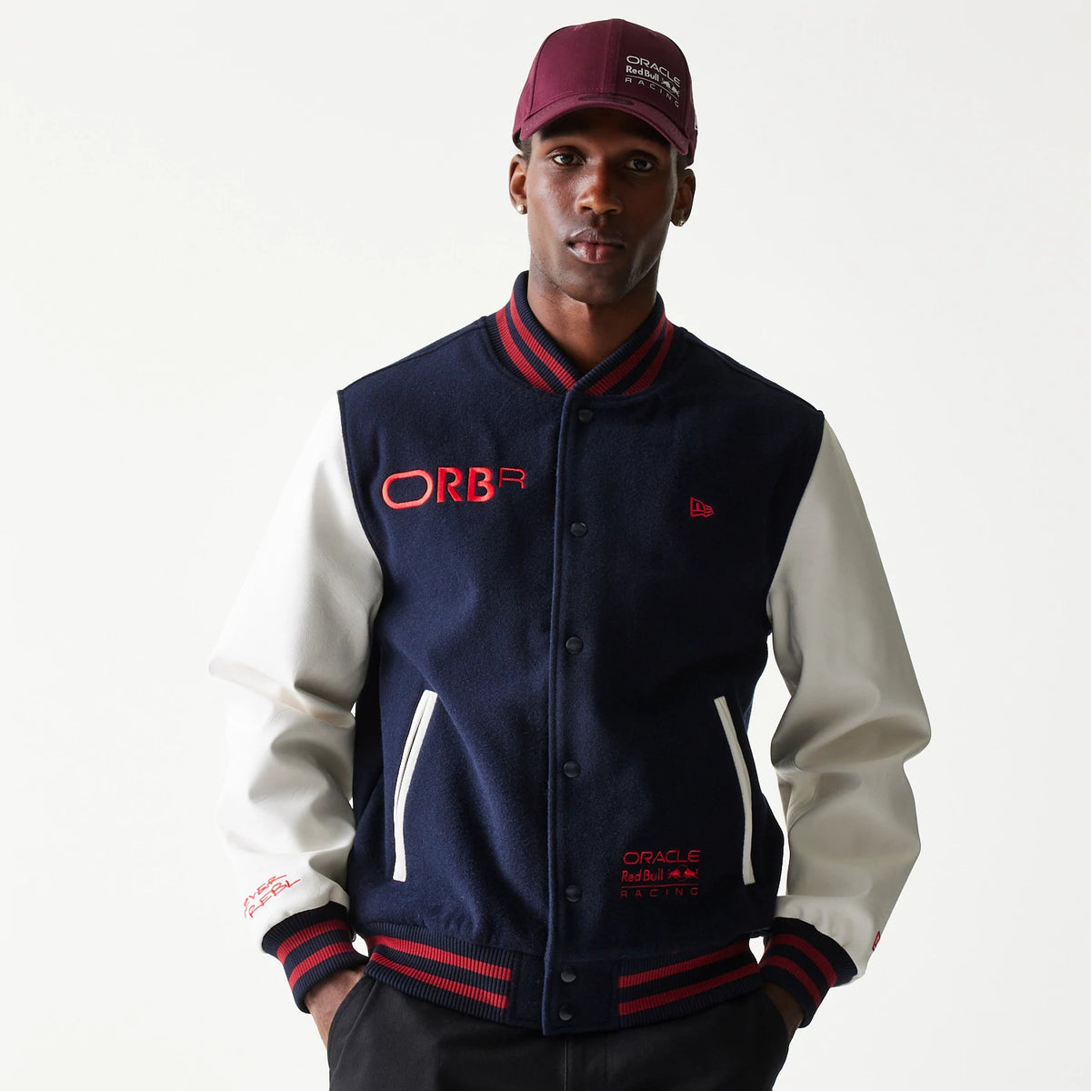 New Era Red Bull Racing Dark Blue Varsity Jacket, front view showing the dark blue body with white sleeves, red lettering on the chest and sleeve, button closure, striped collar, cuffs, hem, and a raised New Era logo on the left chest.