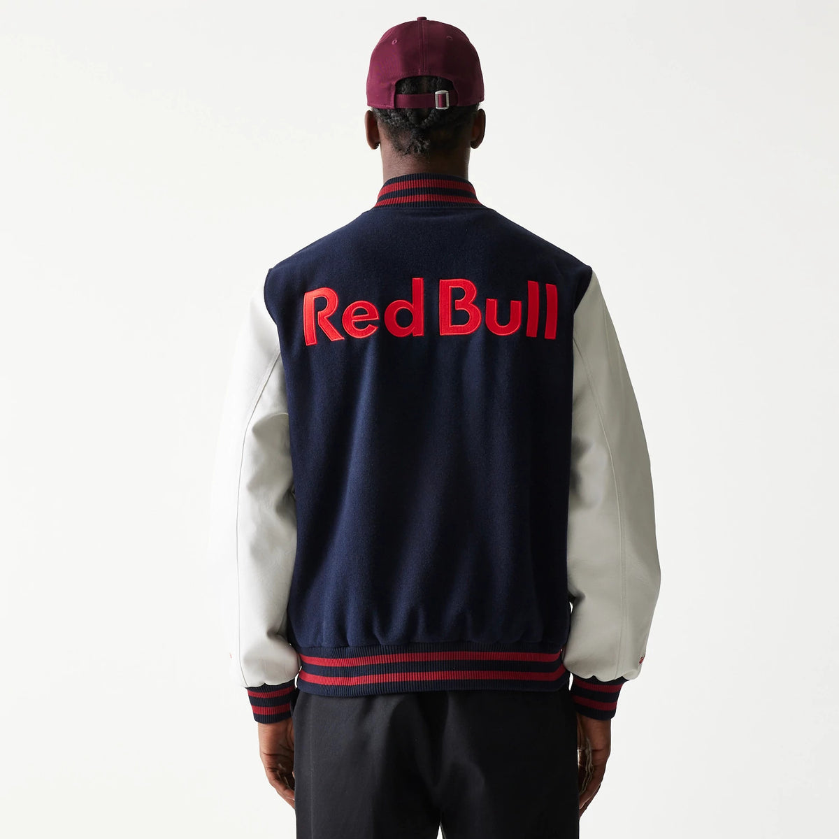New Era Red Bull Racing Dark Blue Varsity Jacket, rear view showing large red embossed Red Bull text across the back of the dark blue body, white sleeves, and a striped red and blue collar and hem, worn by a person in a purple cap.