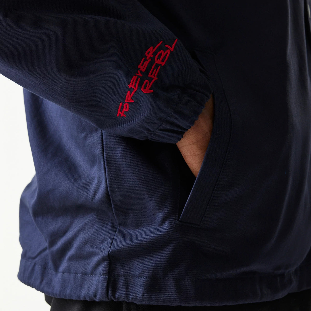 New Era Red Bull Racing Dark Blue Oversized Coach Jacket, side view showing the deep blue fabric with a red embroidered Forever Rebel text on the sleeve above an elasticated cuff and a hand partially tucked into the side pocket.
