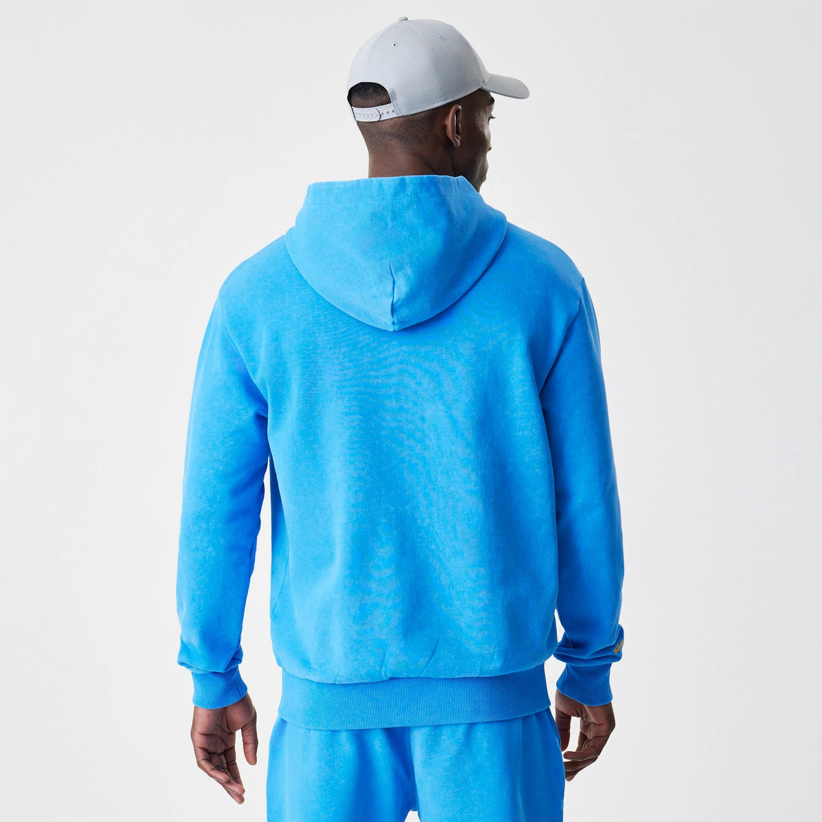 New Era Red Bull Racing Blue Oversized Full Zip Hoodie, back view on a model showing the relaxed hood and ribbed cuffs against the bright blue fabric, paired with matching blue trousers and a light grey cap worn forwards.