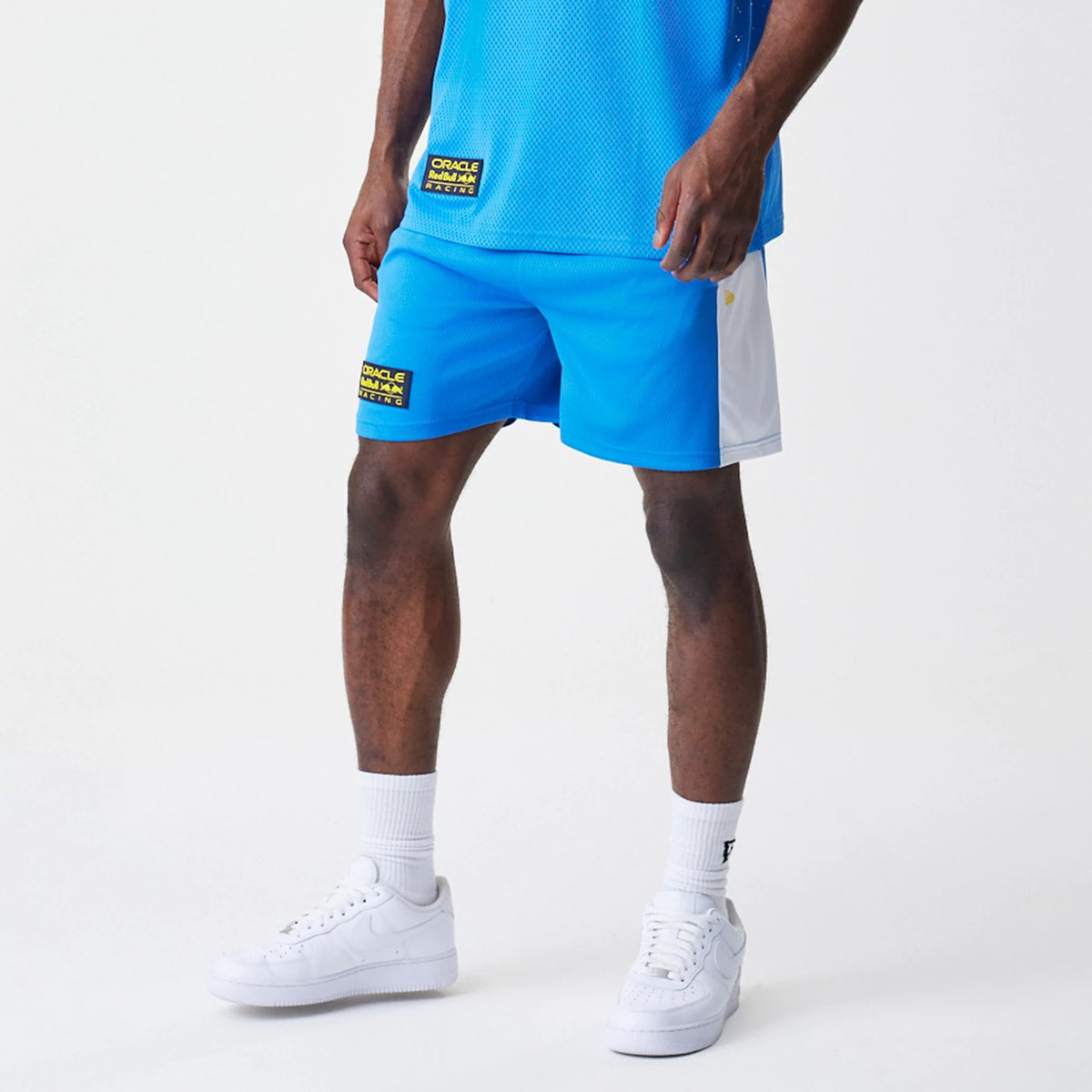 New Era Red Bull Racing Graphic Mesh Blue Shorts, on-model front view showing blue shorts with a white side panel, an embroidered Red Bull Racing logo on the front left, combined with white trainers and socks.