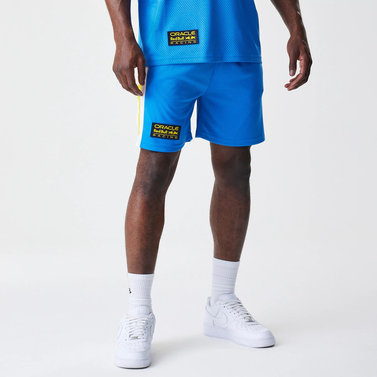 New Era Red Bull Racing Graphic Mesh Blue Shorts, front view on a model styled with a blue top, showing the black and yellow Oracle Red Bull Racing patch on the lower left leg and a yellow stripe running down the side.