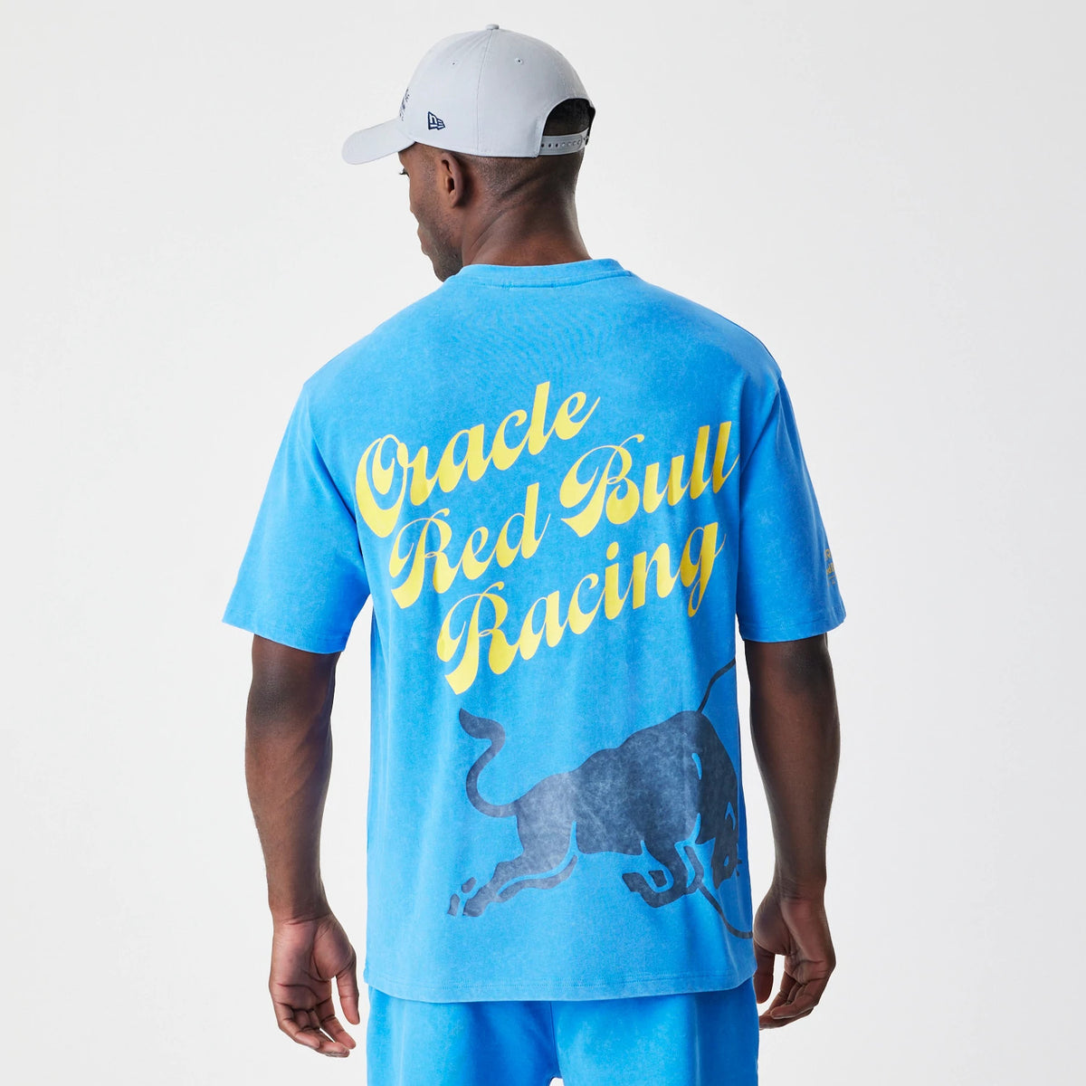 New Era Red Bull Racing Back Graphic Blue Oversized T-Shirt, back view on a model with a grey cap, featuring large yellow Oracle Red Bull Racing lettering and a dark blue bull graphic across the back.