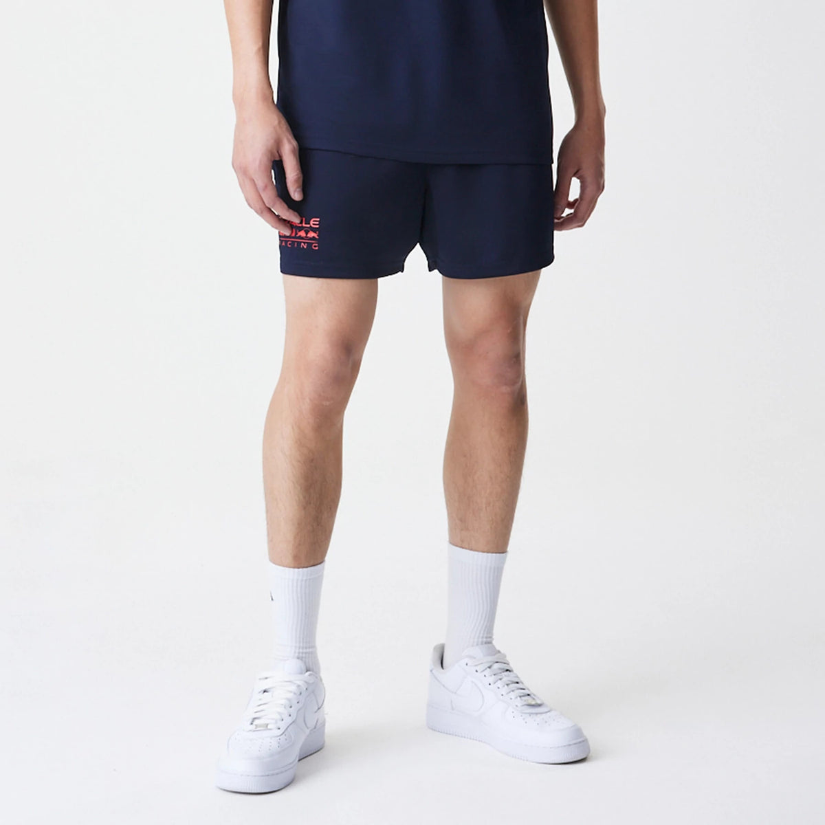 New Era Red Bull Racing Graphic Dark Blue Football Shorts, front view on-model displaying a small red printed Red Bull Racing logo on the left thigh, styled with a matching dark blue top and white trainers for a sporty look.