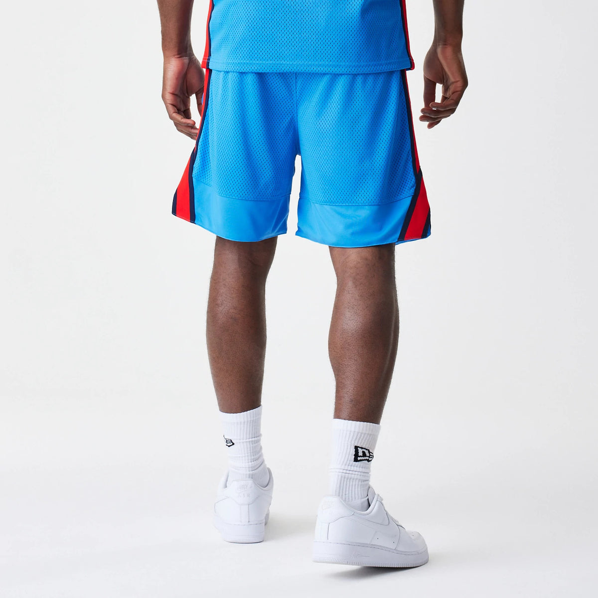 New Era Red Bull Racing Basketball Mesh Blue Shorts, back view on a model showing the blue mesh fabric with red side panels, reaching just above the knee and styled with white trainers and socks featuring a embroidered New Era logo.