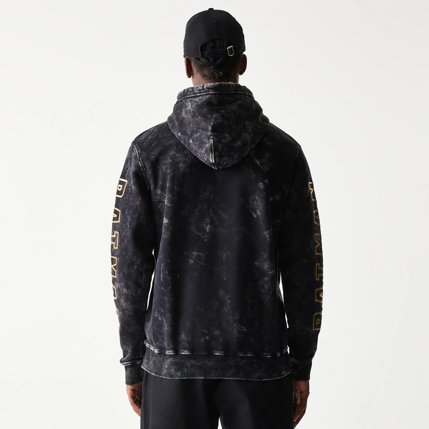New Era Batman 1989 Black Pullover Hoodie, back view on a model with a black cap, showing a faded grey pattern across the hoodie and large Batman lettering in gold down each sleeve.