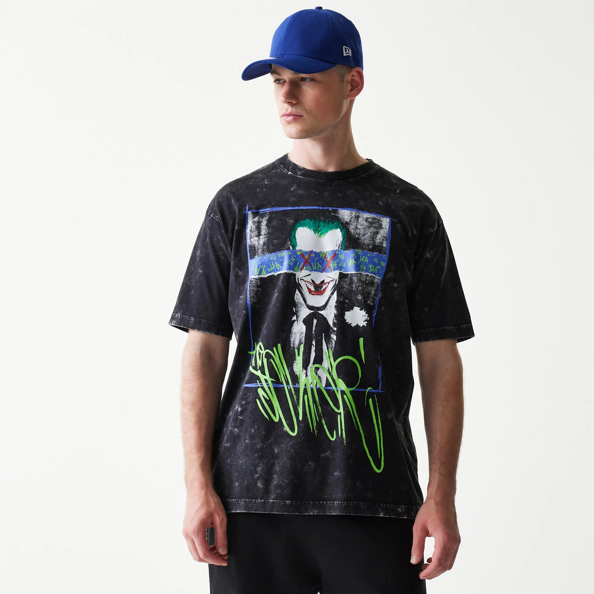 New Era Joker Batman Black T-Shirt, front view on a model showing a graphic of the Joker with colourful accents on the eyes and green lettering below, paired with a plain blue cap.
