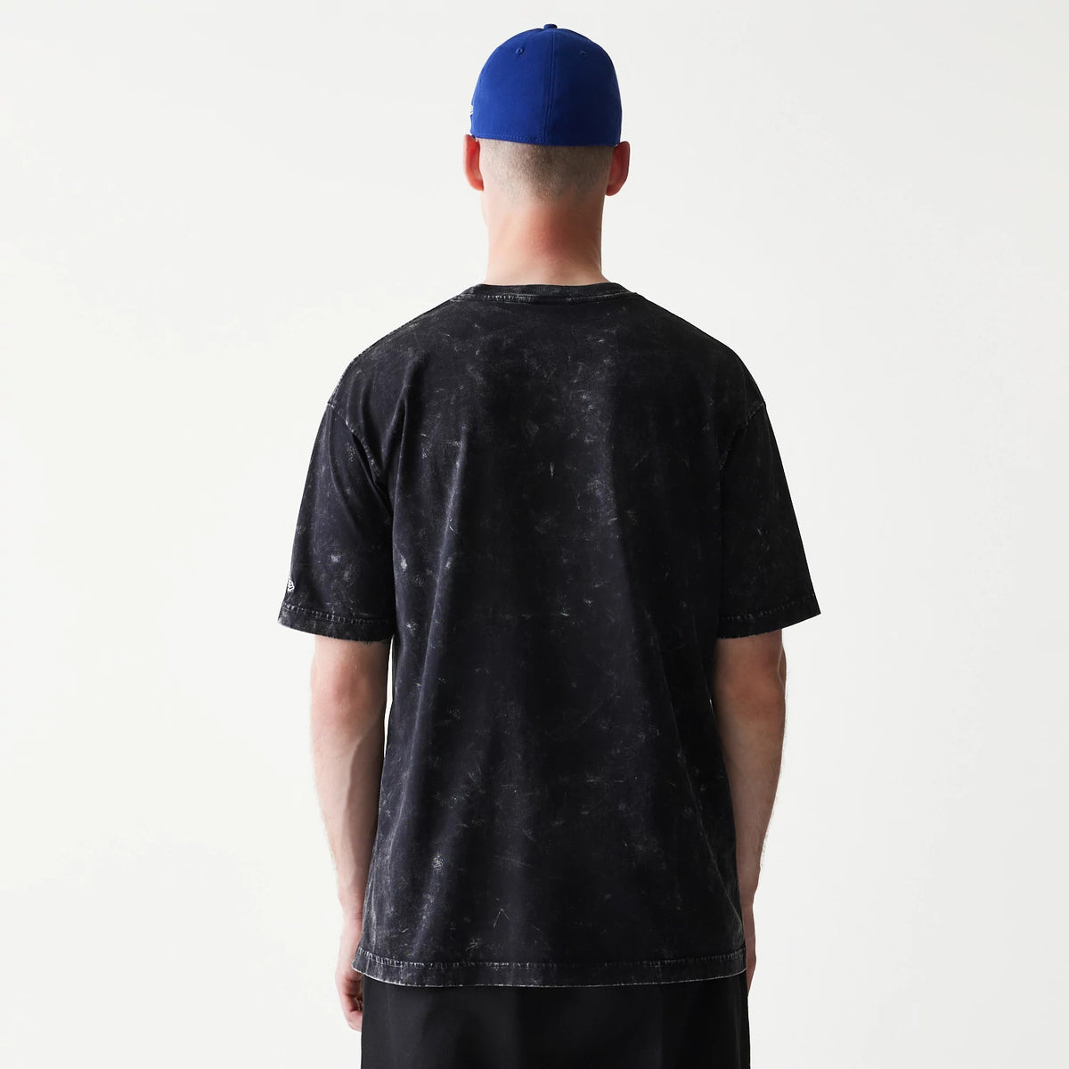 New Era Joker Batman Black T-Shirt, back view on-model showing the black speckled pattern on the fabric, and a small embroidered New Era logo on the left sleeve, paired with a plain blue cap.