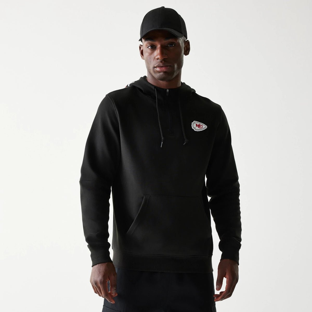 New Era Kansas City Chiefs NFL 3RD Down Black Pullover Hoodie, front view on-model showing the black hoodie with a drawstring hood, a kangaroo pocket, and an embroidered Kansas City Chiefs logo on the left chest, paired with a plain black cap.