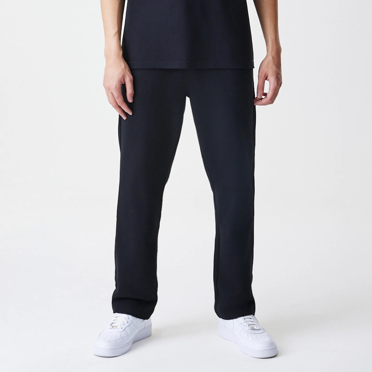 New Era Premium Essential Black Joggers, front view on a model paired with white trainers, featuring a relaxed fit and elastic waistband, showcasing simple and minimalist design in deep black fabric.