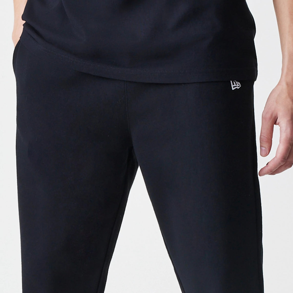 New Era Premium Essential Black Joggers, front view showing the white New Era flag logo embroidered on the upper left pocket area, with a clean black fabric and a relaxed fit around the waist and thighs.