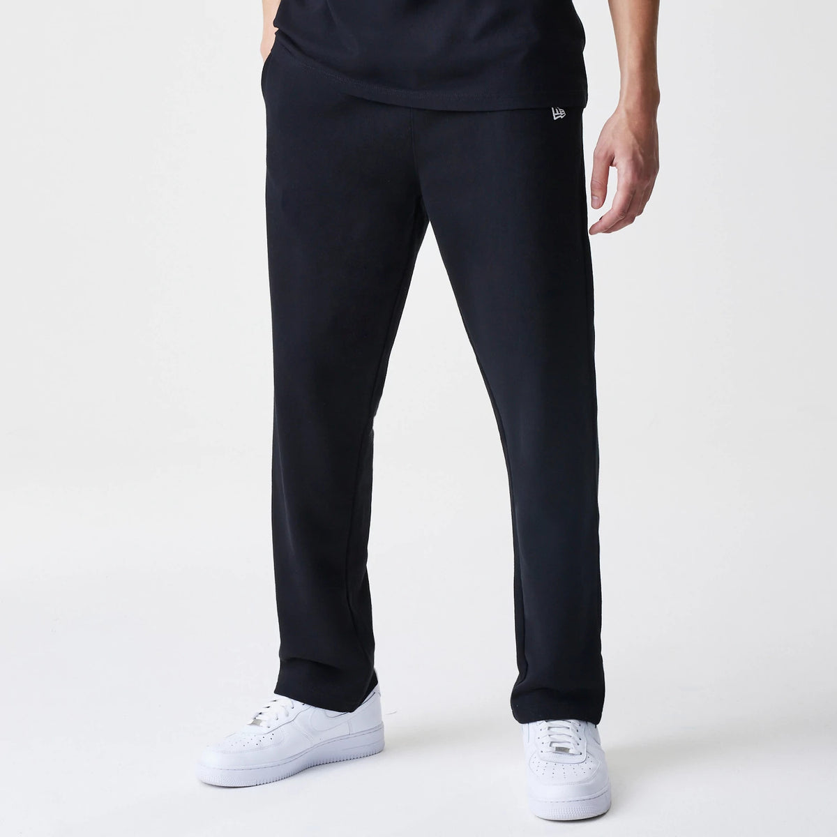 New Era Premium Essential Black Joggers, front view on a model with a white New Era flag logo embroidered on the left thigh, paired with a black top and white trainers for a relaxed casual look.
