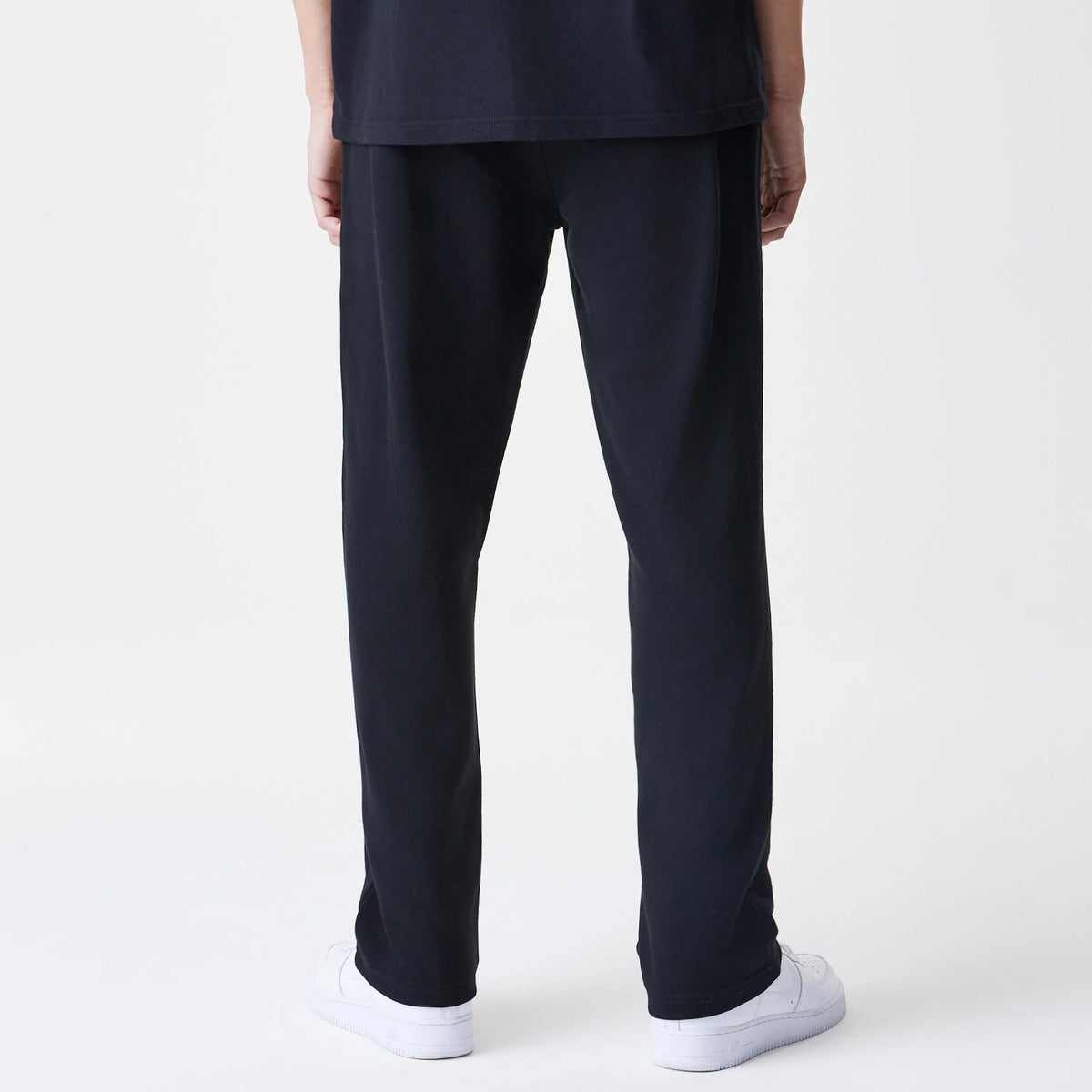 New Era Premium Essential Black Joggers, rear view showing straight leg fit and stitching on a model with a black t-shirt and white trainers, highlighting the simplicity and comfortable design of the joggers.