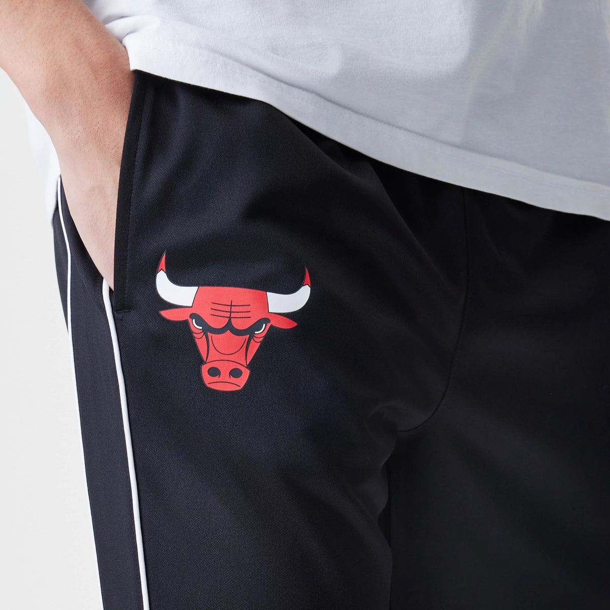 New Era Chicago Bulls NBA Slim Black Track Pants, close-up view showing the red Chicago Bulls logo on the black fabric near the left pocket, with white piping running down the seam for a contrast.