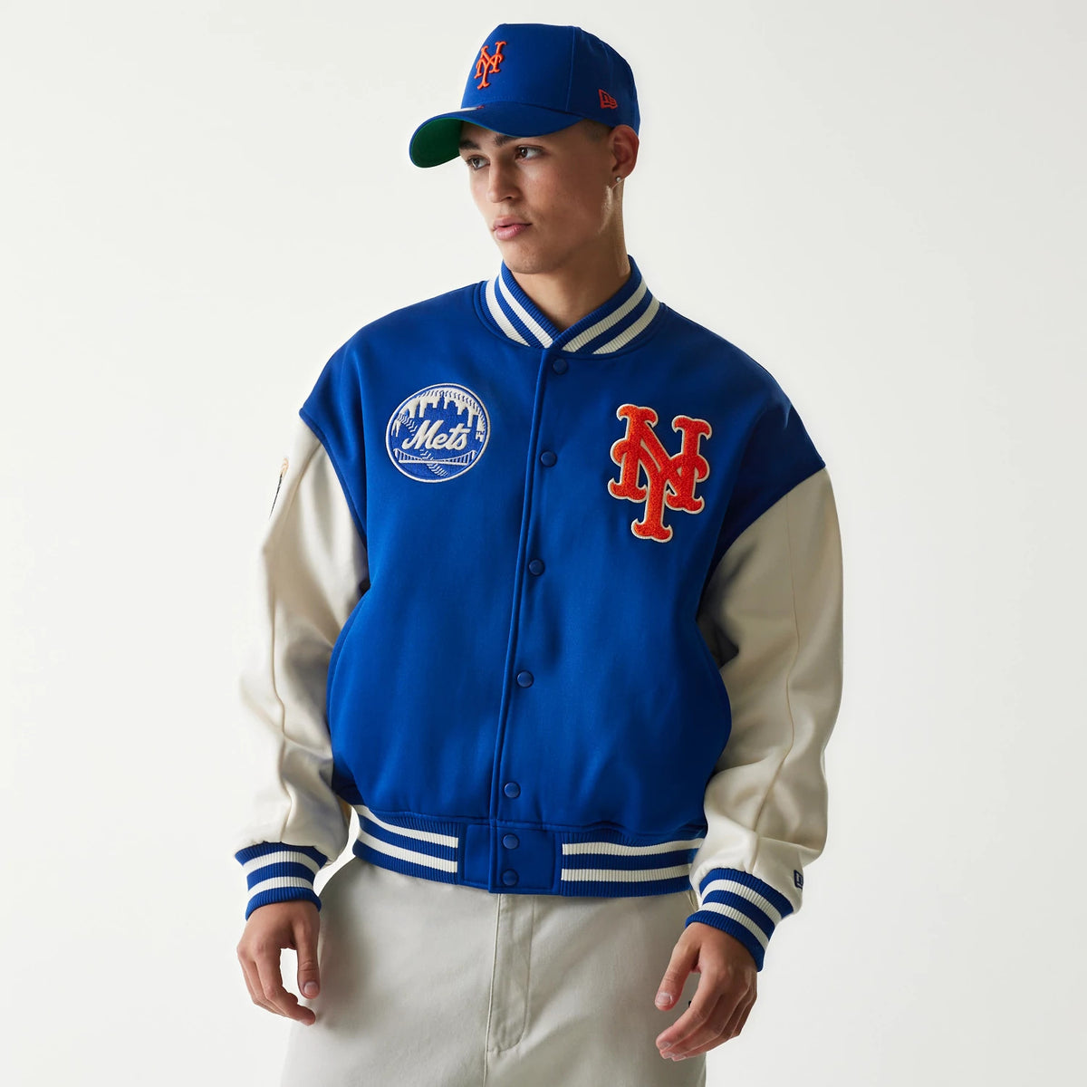 New Era New York Mets MLB Jersey Blue Bomber Jacket, front view on a model wearing the jacket with distinctive orange embroidered NY logo on the chest, matching blue and white striped collar and cuffs, and a Mets patch on the right side.