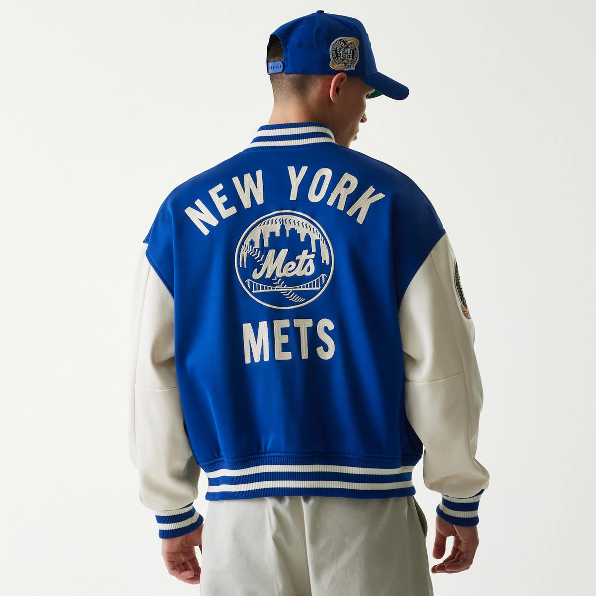 New Era New York Mets MLB Jersey Blue Bomber Jacket, back view on a model showing large white embroidered New York Mets lettering and logo centred on the blue back, with striped cuffs and waistband, paired with a matching blue cap.