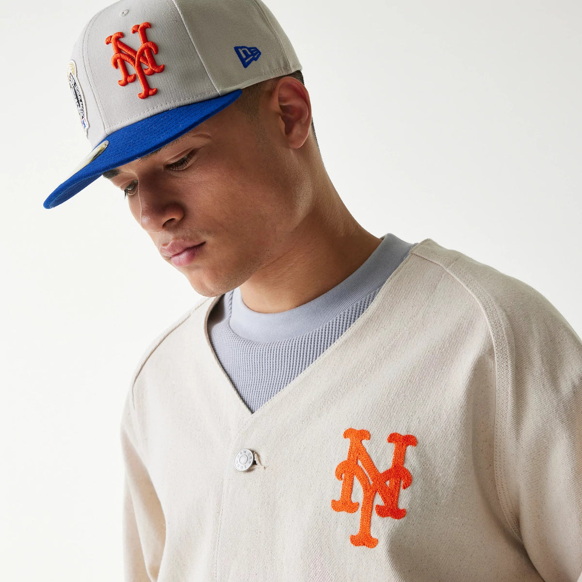 New Era New York Mets MLB Cotton Open Cream Jacket, front view on-model showing the orange embroidered team logo on the left chest, with a cream button closure, styled with a grey top and matching cap.
