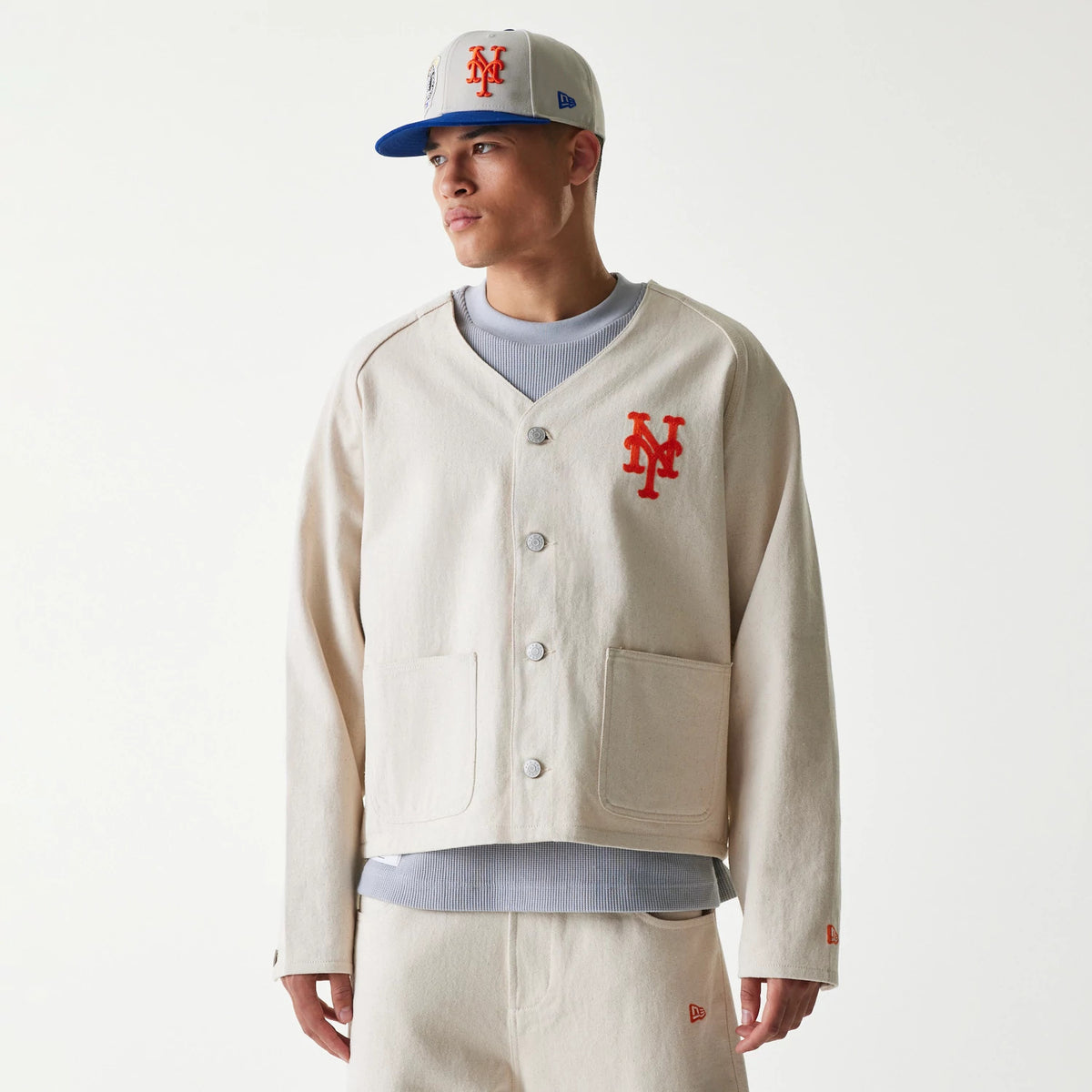 New Era New York Mets MLB Cotton Open Cream Jacket, front view on-model showing a cream jacket with a large orange embroidered NY logo on the chest, button closure, two front pockets, and a small New Era logo on the sleeve.