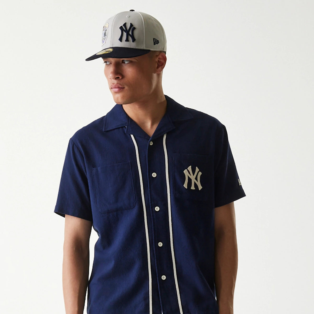 New Era New York Yankees MLB Piping Navy Short Sleeve Shirt, front view on a model paired with a matching navy and white cap, showing the white New York Yankees logo on the left chest and piping along the button placket.