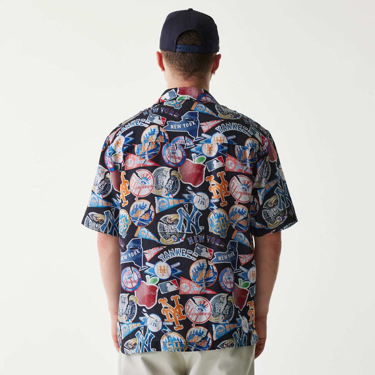 New Era MLB Subway Series All Over Print Navy Short Sleeve Shirt, back view on a model showing a design featuring various New York Yankees and Mets logos, baseball imagery, and city symbols on the navy fabric.